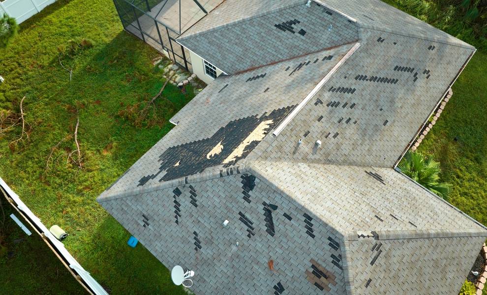 An aerial view of a residential roof showing significant storm damage, with missing and torn shingles exposing sections of the underlying roof surface.