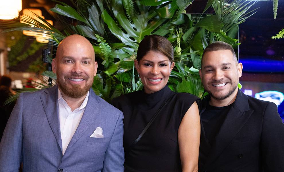 Three people pose together at an evening event, smiling in front of a lush backdrop of tropical greenery.
