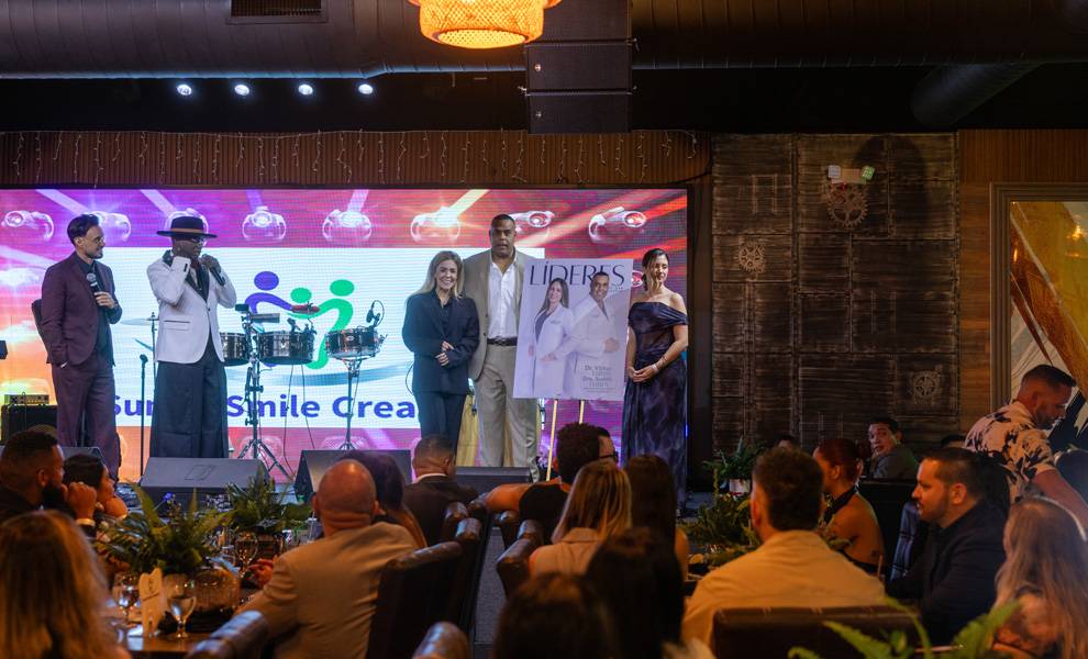 A live event takes place indoors with a stage featuring speakers and performers under warm lighting. A group stands on stage near a large screen displaying “Sunset Smile,” while an audience sits at tables watching the presentation in a lively, upscale venue.