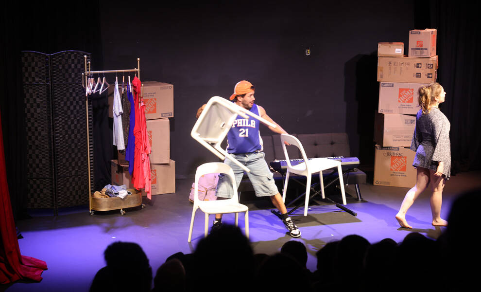 En una representación teatral en vivo titulada «Mi Vida en Alquiler», se observa en el escenario a un hombre —vestido con ropa informal y una camiseta deportiva— levantando enérgicamente una silla en plena escena, mientras una mujer se retira hacia un lateral. La escenografía incluye cajas de mudanza apiladas, un perchero con prendas de vestir y un mobiliario minimalista, lo que sugiere una escena de carácter cómico o dramático centrada en una mudanza o en la vida cotidiana. El público resulta visible, en silueta, en primer plano.