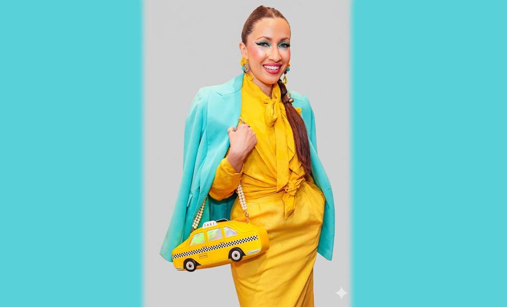 Yli Liriano in a bright yellow dress with a teal blazer draped over her shoulders smiles while holding a novelty yellow taxi-shaped handbag against a light background.