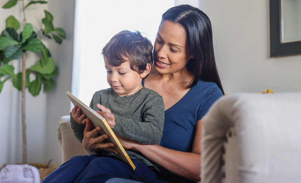 Escena de apoyo familiar de Aetna que muestra a una madre leyendo con su hijo pequeño en casa, mientras miran juntos un libro en una cómoda sala de estar.