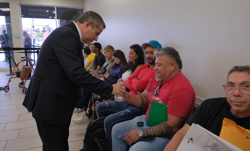 Dariel Fernandez saluda a los residentes en una sala de espera de una oficina de Miami-Dade, estrechando la mano de un visitante mientras otros aguardan con documentos y pertenencias personales.
