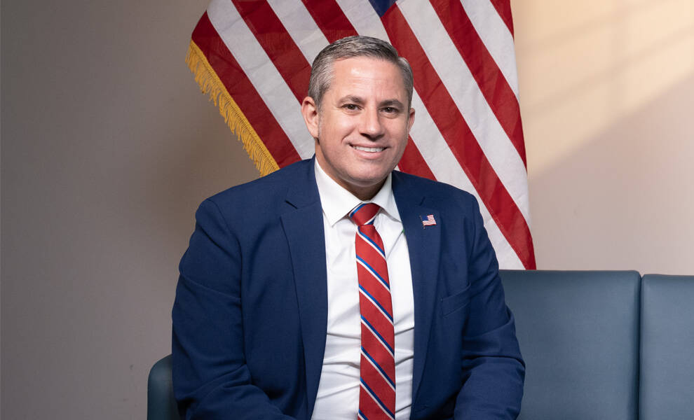El Recaudador de Impuestos del Condado de Miami-Dade, Dariel Fernandez, se sienta frente a una bandera estadounidense, vistiendo un traje azul y una corbata de rayas rojas.