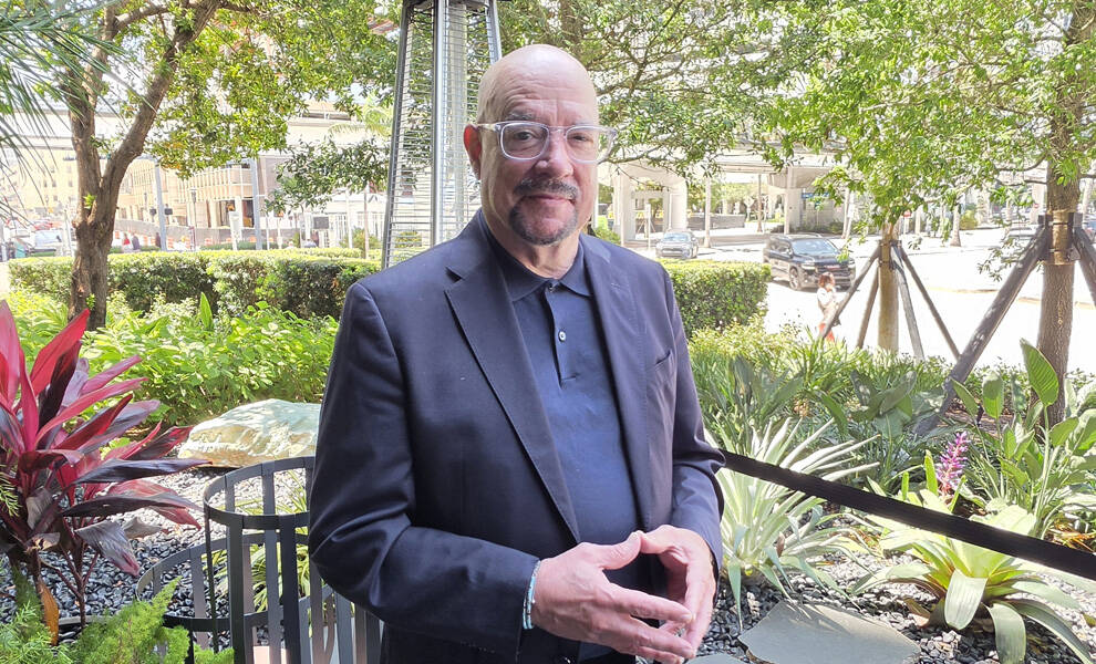 David Whitaker, presidente y director ejecutivo del GMCVB, posa con un traje oscuro en una zona ajardinada con palmeras, vegetación y un calentador de patio visible al fondo.