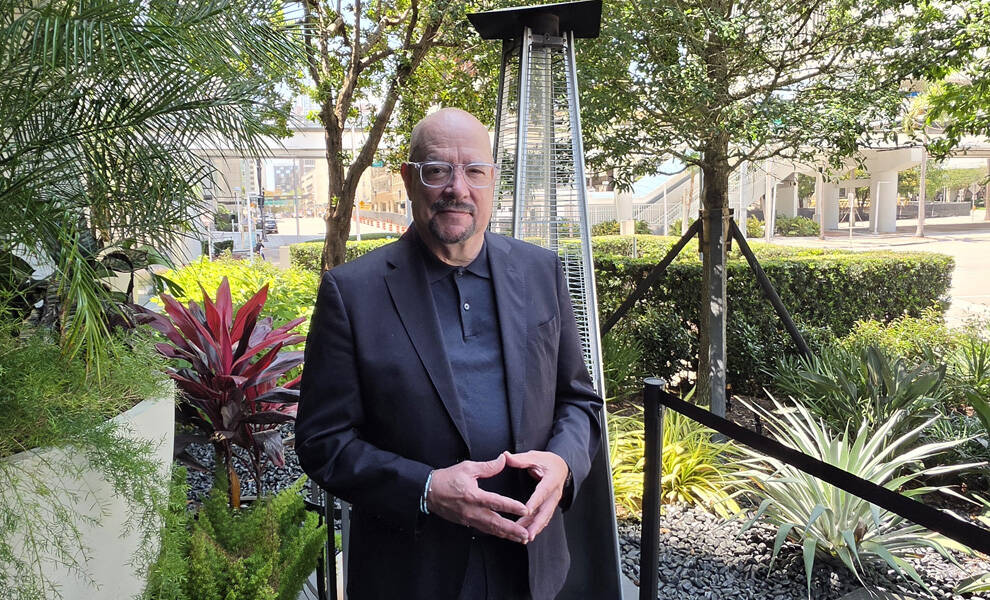 David Whitaker GMCVB President & Chief Executive Officer wearing glasses and a dark blazer stands outdoors in a landscaped courtyard surrounded by tropical plants and trees in an urban setting.
