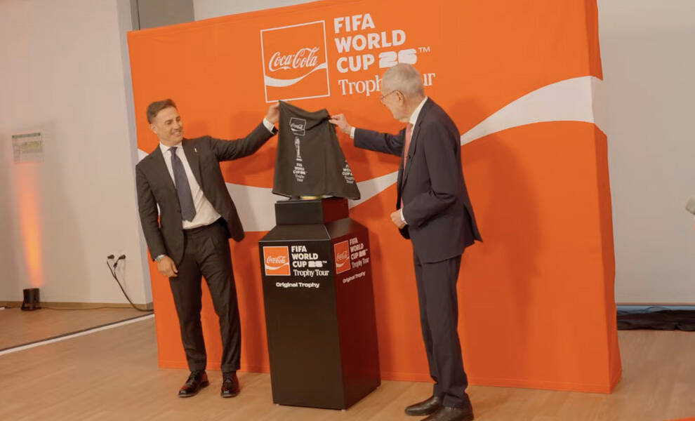 Two men in suits stand beside a display pedestal at a Coca-Cola FIFA World Cup 2026 Trophy Tour event.