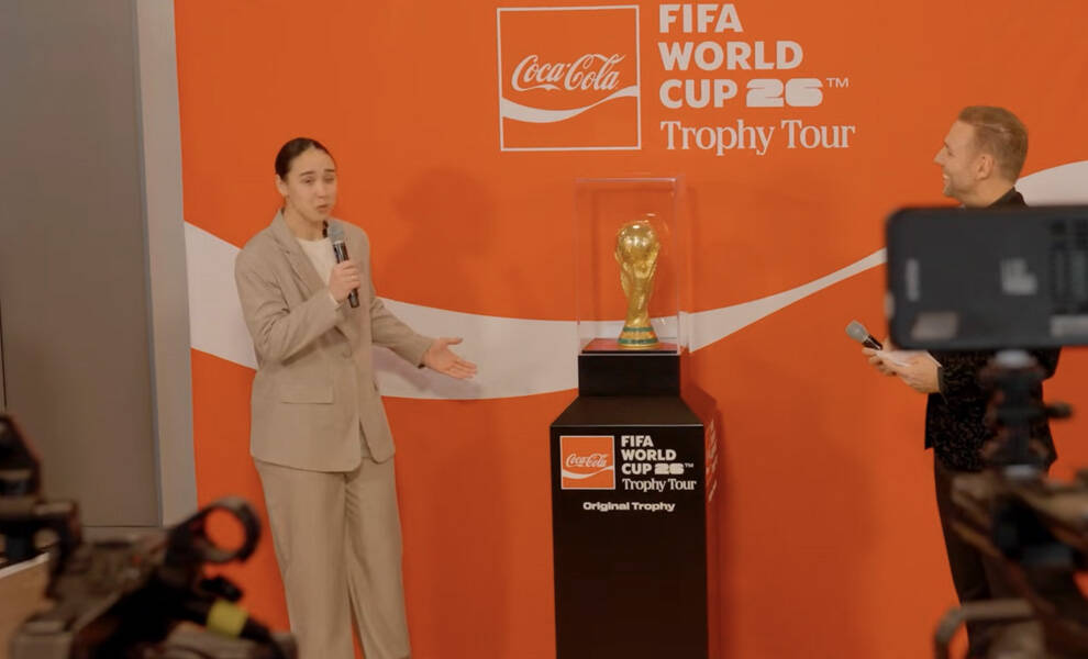 Un orador, vestido con un traje beige, hace un gesto hacia el trofeo de la Copa Mundial de la FIFA, exhibido en una vitrina de cristal, durante un evento de la Gira del Trofeo de la Copa Mundial de la FIFA 2026 de Coca-Cola.