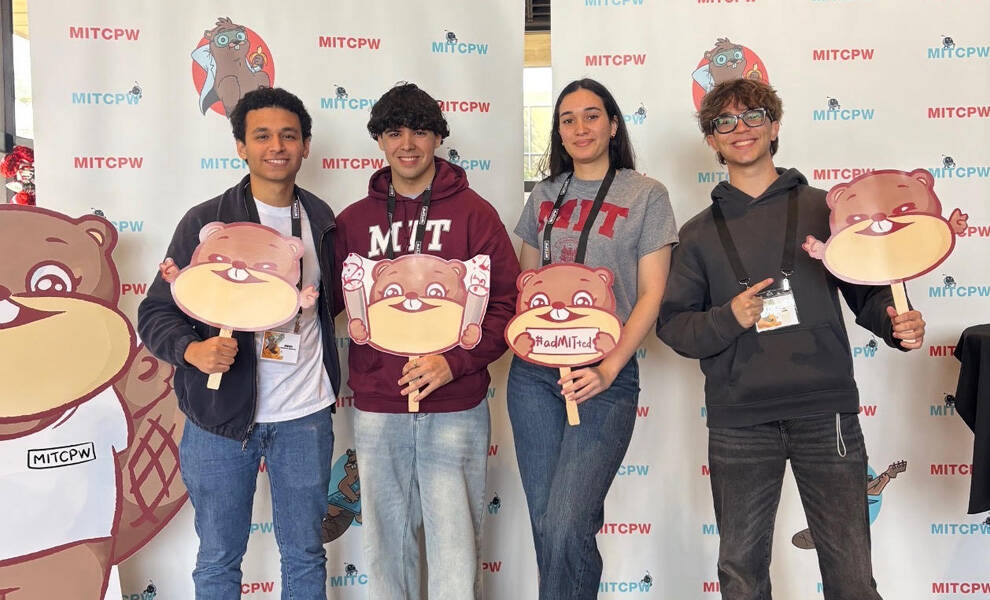 Cuatro estudiantes posan frente a un telón de fondo de un evento del MITCPW, sosteniendo carteles con dibujos animados de castores y sonriendo durante una conferencia estudiantil centrada en la academia y la tecnología.