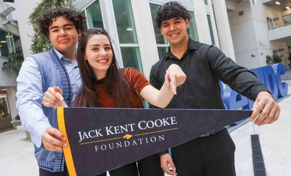 Tres estudiantes permanecen juntos en un campus universitario, sosteniendo un banderín de la Jack Kent Cooke Foundation y celebrando los logros académicos y las oportunidades de becas.