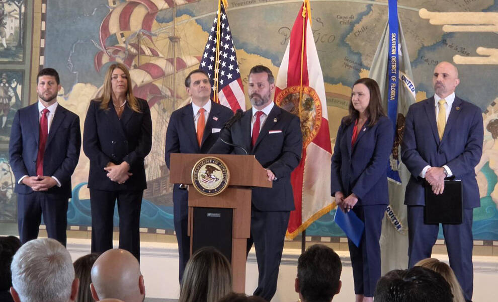 Funcionarios gubernamentales y representantes de las fuerzas del orden se sitúan detrás de un podio durante una conferencia de prensa, con las banderas de Estados Unidos y del estado de Florida exhibidas al fondo.