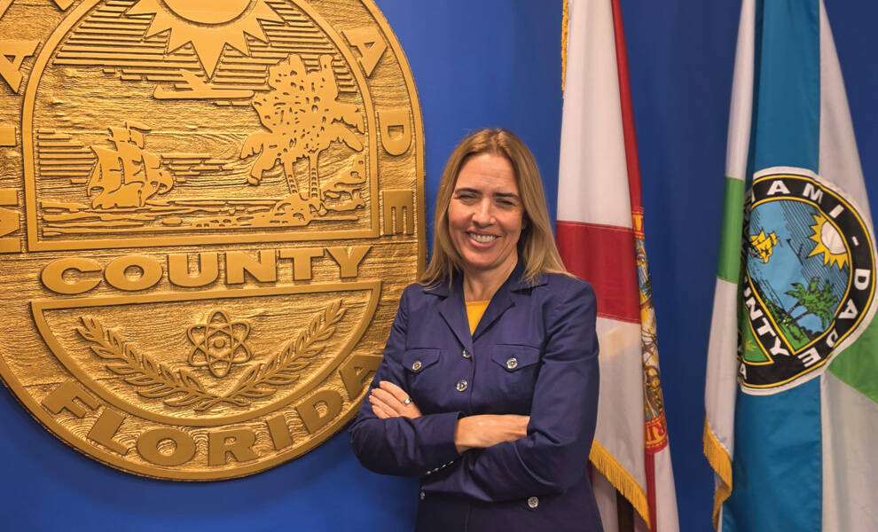 Raquel Regalado posa sonriente, con los brazos cruzados, frente al sello del condado de Miami-Dade, con las banderas de Florida y de Miami-Dade exhibidas al fondo.