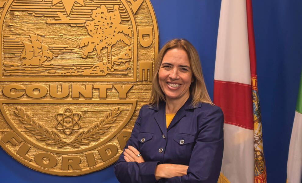 Raquel Regalado stands smiling with folded arms in front of the Miami-Dade County seal, with Florida and Miami-Dade flags displayed in the background.
