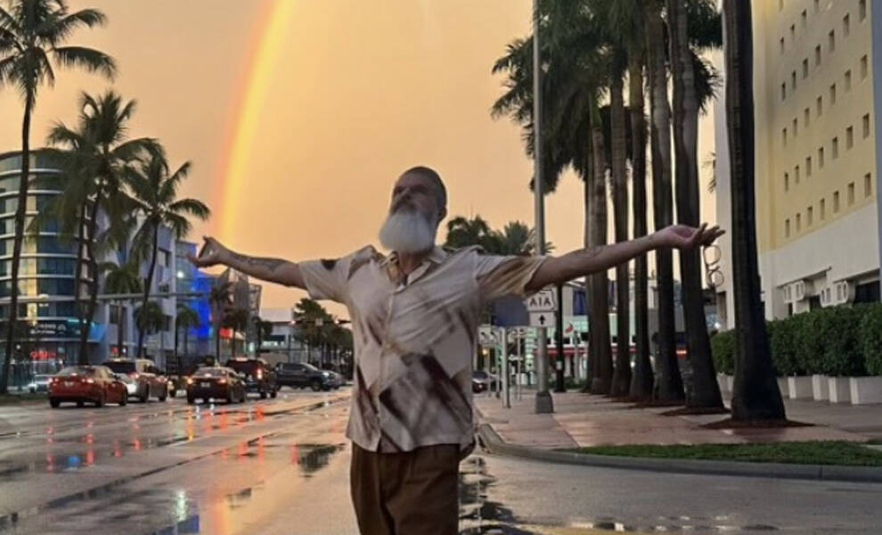 Sexton Garcia stands in the middle of a rain-soaked street with his arms outstretched as a bright rainbow arches behind him. Palm trees line both sides of the road, and reflections from the wet pavement create a colorful glow against the evening sky.