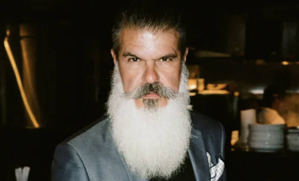 Sexton Garcia with a long white beard and styled mustache poses in a dark setting while wearing a gray suit jacket and white pocket square.