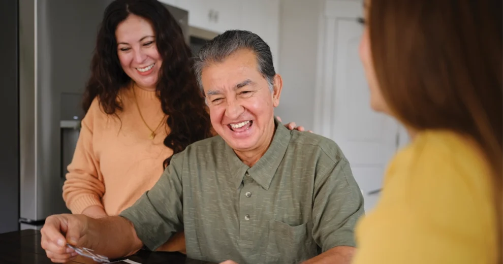An older man with Alzheimer's disease smiles warmly while seated at a table in a bright home setting, holding a pair of eyeglasses. A woman stands beside him with her hand on his shoulder, smiling as they share a pleasant moment.