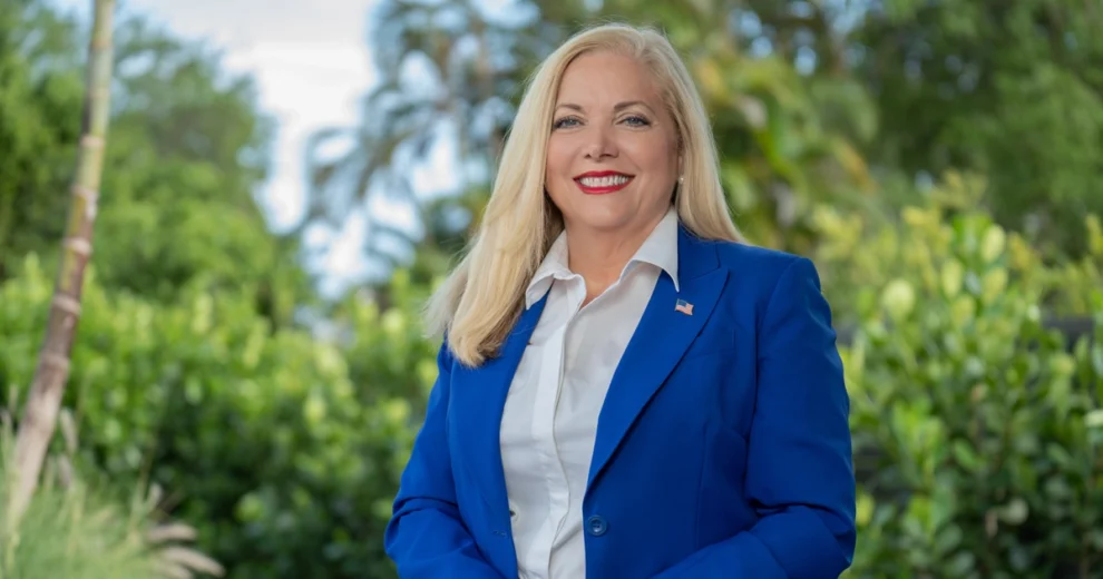 La Supervisora ​​de Elecciones del condado de Miami-Dade, Alina García —de largo cabello rubio— sonríe de pie al aire libre, en un entorno tropical ajardinado.