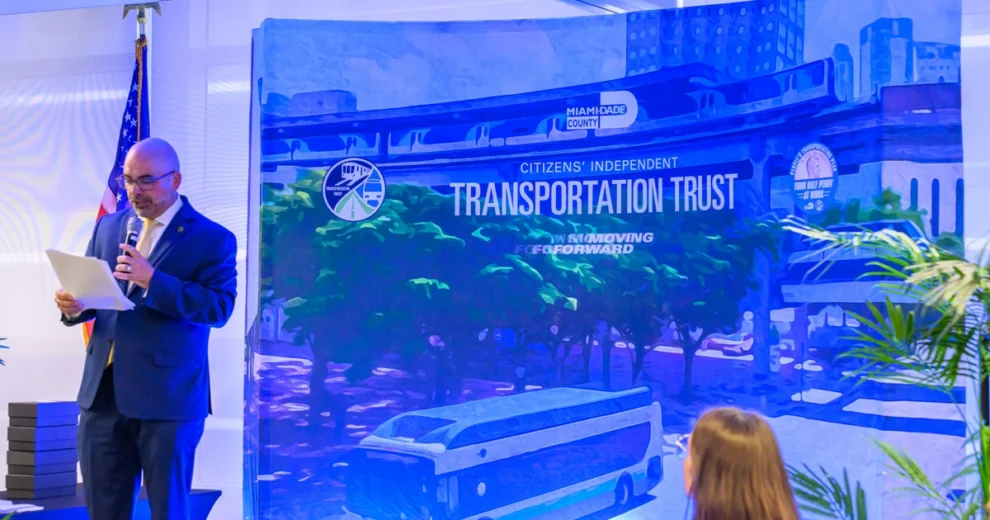 Un hombre con traje azul marino y corbata amarilla está de pie en un escenario, hablando por un micrófono mientras lee una hoja de papel durante un evento del Citizens’ Independent Transportation Trust.