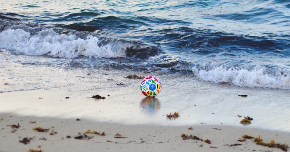 An international soccer ball stands still on the sand as a group of people where enjoying a game of soccer on the beach.