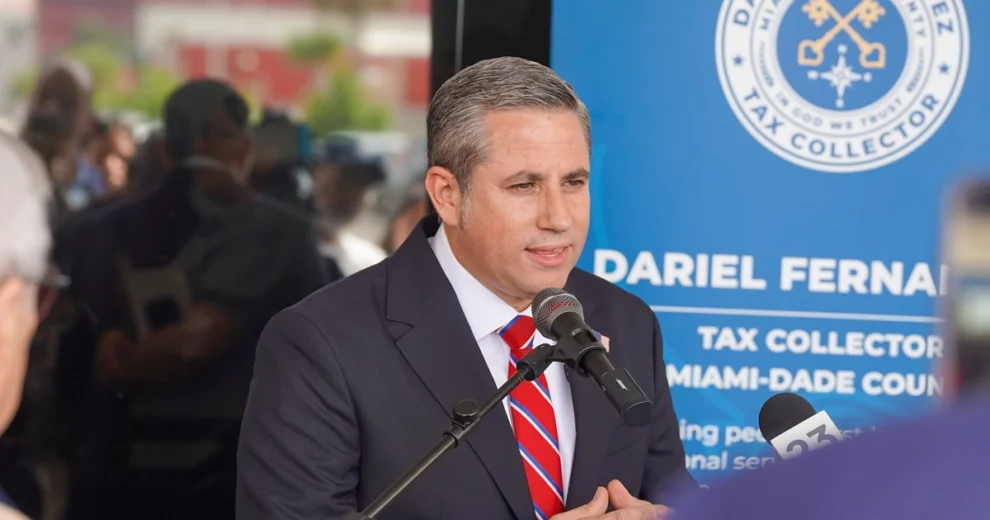 Miami-Dade County Tax Collector Dariel Fernandez speaks at a public event while standing behind a microphone. He is wearing a dark suit, white shirt, and red-and-blue striped tie.