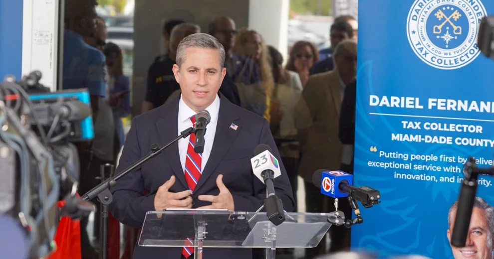Miami-Dade County Tax Collector Dariel Fernandez speaks at a podium during a press event, with microphones and cameras in the foreground and a large branded banner beside him.