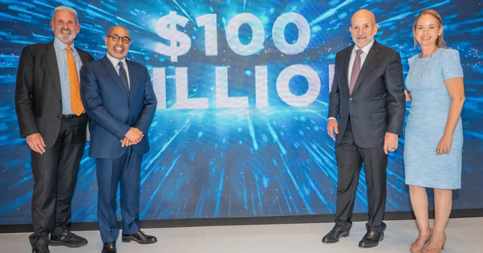 Cuatro personas vestidas de negocios posan frente a una gran pantalla digital que muestra "100 millones de dólares" en letras blancas y negritas sobre un fondo azul.