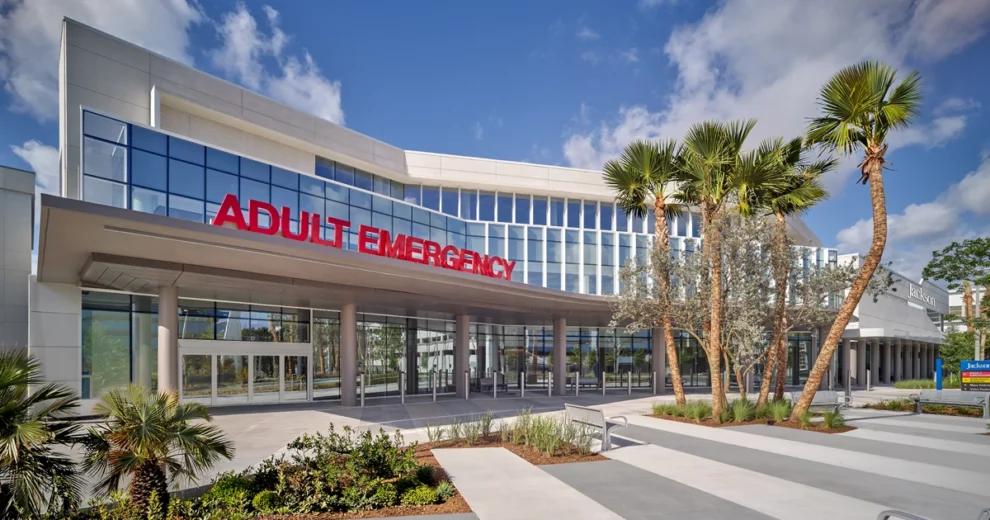 Vista exterior de un moderno edificio del Jackson Memorial Hospital que presenta una amplia entrada cubierta, señalizada con las palabras "Adult Emergency" en letras de color rojo brillante.