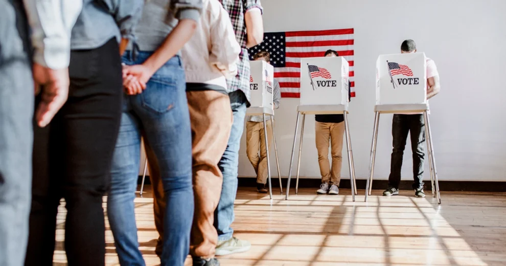 Voters stand in line at an early voting polling place while others cast ballots in private voting booths. An American flag hangs on the wall behind the booths, highlighting the civic process of voting and participation in elections.