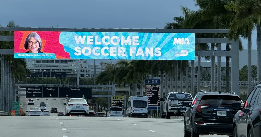 A large digital welcome banner spans the roadway entrance to Miami International Airport, displaying the message &ldquo;Welcome Soccer Fans&rdquo; alongside a portrait of Mayor Daniella Levine Cava.