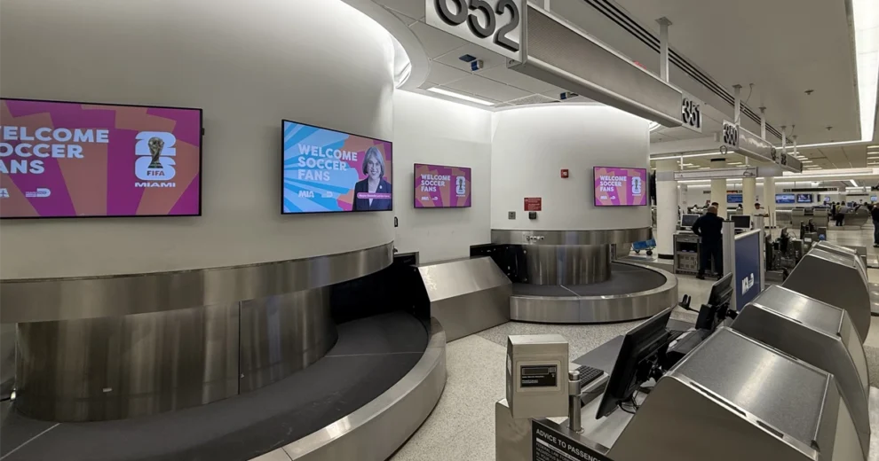 Inside Miami International Airport’s baggage claim area, multiple digital screens display “Welcome Soccer Fans” messages featuring FIFA World Cup branding and Mayor Daniella Levine Cava.