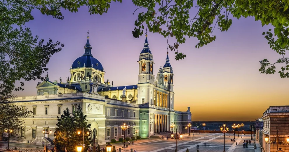 Illuminated historic cathedral and palace complex in Madrid at dusk, featuring a large blue-domed roof, ornate towers, and classical architecture.