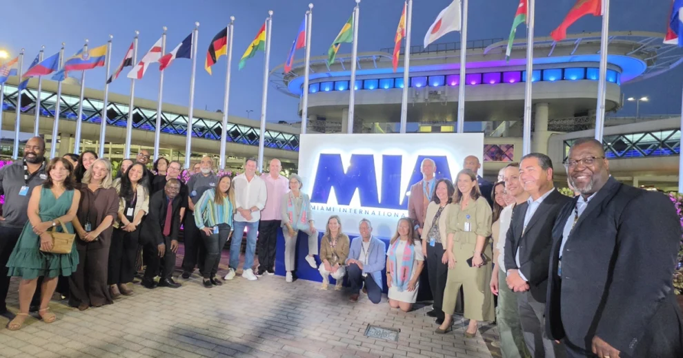 A large group of community leaders, officials, and event organizers pose for a photo outside Miami International Airport (MIA) at dusk. The group stands around an illuminated MIA sign, with international flags displayed on tall flagpoles behind them.