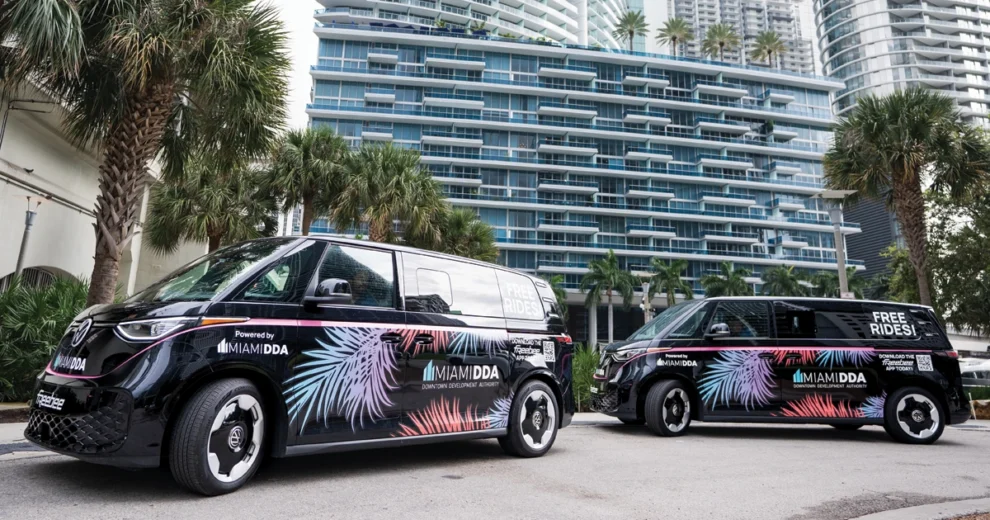 Dos furgonetas eléctricas de transporte, rotuladas y operadas por la Autoridad de Desarrollo del Centro de Miami (Miami Downtown Development Authority), están estacionadas en una calle del centro de la ciudad flanqueada por palmeras y torres residenciales de gran altura. Los vehículos negros lucen coloridos gráficos tropicales y mensajes que anuncian "viajes gratuitos", destacando así un servicio de transporte disponible en la zona céntrica.