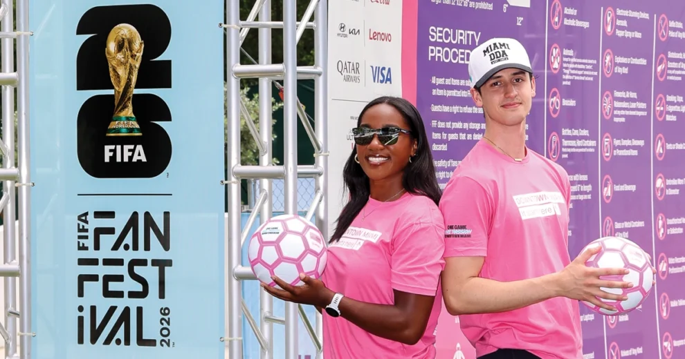 Two event ambassadors wearing bright pink shirts pose with soccer balls beside a FIFA Fan Festival 2026 display.