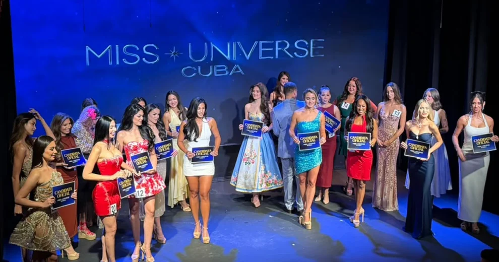 Contestants for Miss Universe Cuba stand on a theater stage during an official selection event. The women are dressed in a variety of formal gowns, cocktail dresses, and eveningwear while holding signs that read “Candidata Oficial.”
