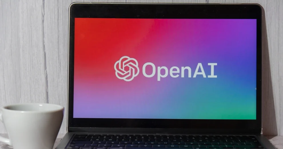 Una computadora portátil descansa sobre un escritorio, junto a una taza de café blanca, mostrando el logotipo y el nombre de OpenAI en una pantalla con un degradado colorido. La imagen representa la tecnología de inteligencia artificial, la innovación digital y la marca OpenAI.