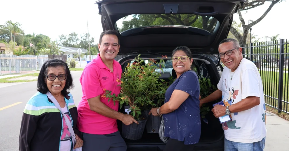 El comisionado del Distrito 4, Ralph "Rafael" Rosado, posa sonriente junto a tres miembros de la comunidad y el maletero abierto de un automóvil lleno de plantas, durante un evento comunitario al aire libre.