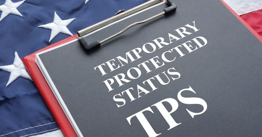Primer plano de una carpeta con pinza roja que sostiene un documento titulado "Estatus de Protección Temporal (TPS)", colocado sobre una bandera de los Estados Unidos. La imagen simboliza la política migratoria, las protecciones humanitarias y la documentación legal relacionada con el Estatus de Protección Temporal en los Estados Unidos.