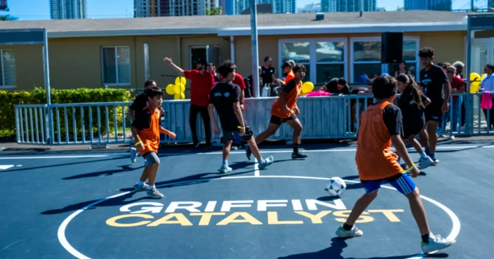 Children and teenagers play a fast-paced soccer match on a Griffin Catalyst-branded outdoor court.
