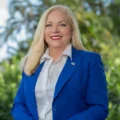 Miami-Dade County Supervisor of Elections Alina Garcia with long blonde hair smiles while standing outdoors in a landscaped tropical setting.