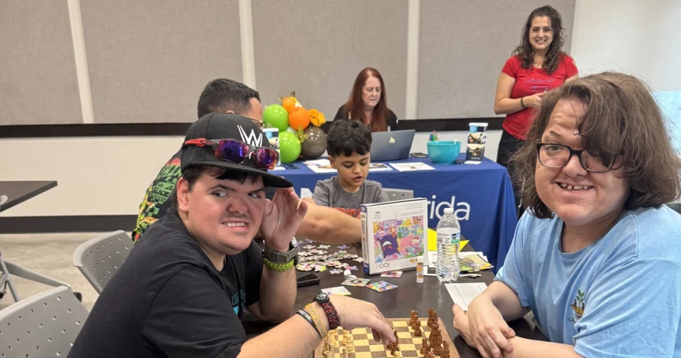 Cumbre de Líderes de ACES con un Destino para Conectar a Familias del Sur de Florida con Recursos para Discapacitados Antes del Año Escolar 1 Dos participantes sonrientes están sentados a una mesa con un tablero de ajedrez durante un evento comunitario de ACES. Otros asistentes, al fondo, realizan rompecabezas, manualidades y diversas actividades, mientras los voluntarios ayudan a los participantes por toda la sala.