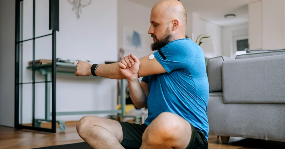 Un afiliado de Aetna está sentado en una esterilla de yoga en la sala de estar y se estira un brazo; lleva puesto un monitor continuo de glucosa en la parte superior del brazo y un reloj inteligente en la muñeca, lo que pone de relieve el control de la diabetes y un estilo de vida saludable en el hogar.