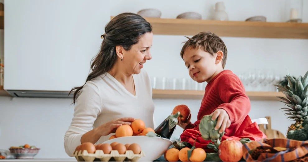 Un padre o una madre y su hijo pequeño, sonrientes, preparan comida saludable juntos en una cocina luminosa. Frutas frescas, verduras y huevos están dispuestos sobre la encimera mientras seleccionan los alimentos y disfrutan cocinando en familia.