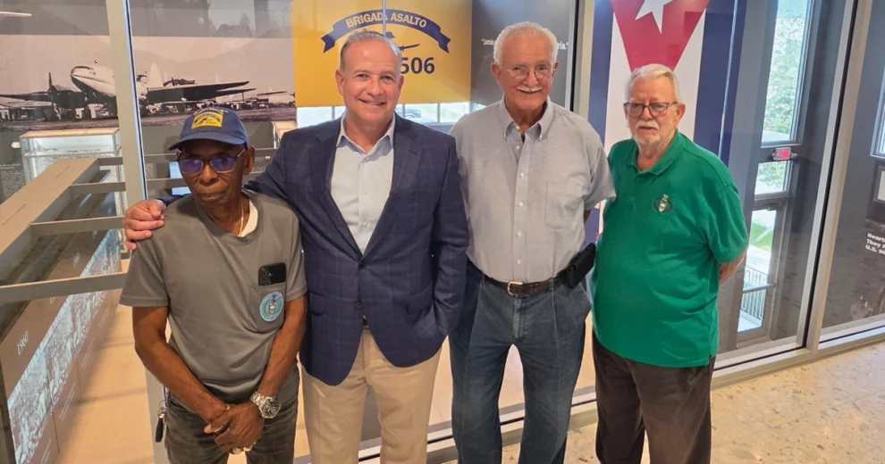 De la traición a la resiliencia: el Museo de Bahía de Cochinos preserva el legado vivo de la Brigada 2506 1 Cuatro hombres posan juntos en el interior del Museo de Bahía de Cochinos, frente a exposiciones históricas, una bandera cubana y un letrero de la Brigada de Asalto 2506. Eduardo Zayas-Bazán, que viste una camisa azul claro abotonada, está de pie junto a un hombre con americana azul y otros dos hombres vestidos de manera informal con una camiseta gris y un polo verde. El grupo posa para una foto conmemorativa en la zona de exposición del museo.