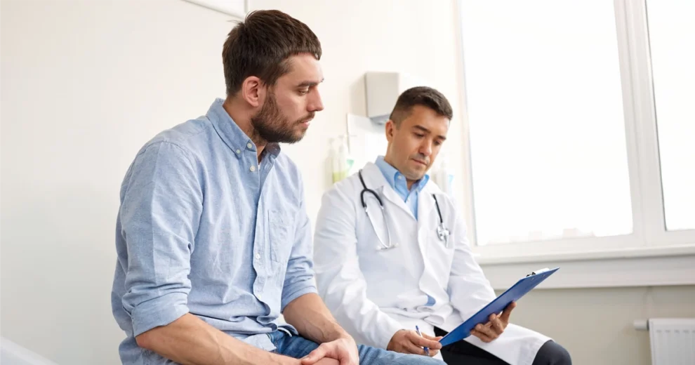 Un médico de Care Resource revisa la información médica de un paciente en una carpeta con sujetapapeles durante una consulta. El paciente permanece sentado atentamente en la sala de exploración mientras habla sobre su atención médica con el doctor.