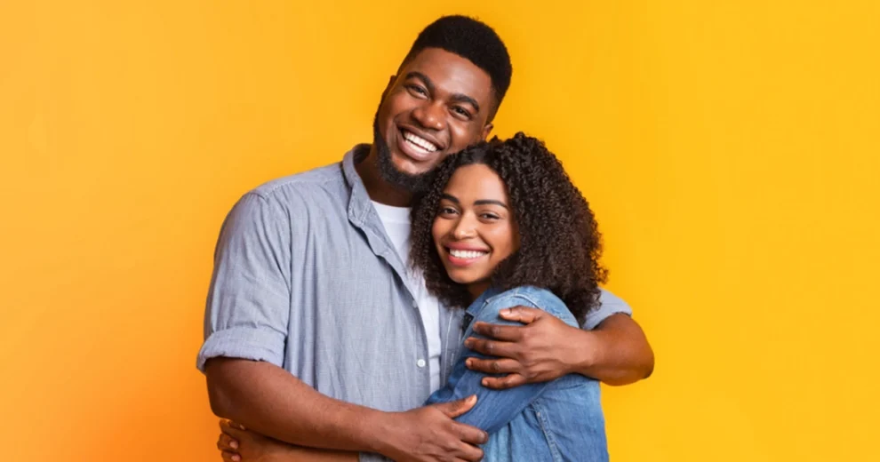 A smiling couple embraces against a bright yellow background. The taller person wraps an arm around their partner as both smile warmly at the camera, illustrating love, support, acceptance, and human connection.