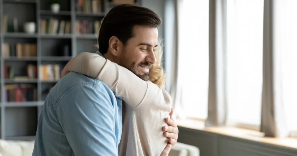 A person embraces another individual in a bright indoor setting near a window. The comforting hug conveys empathy, reassurance, and emotional support, reinforcing the article's message about challenging HIV stigma.