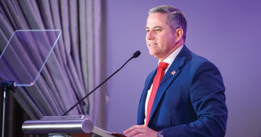 El recaudador de impuestos del condado de Miami-Dade, Dariel Fernandez, habla desde un podio durante un evento formal. Viste traje azul, camisa blanca de vestir, corbata roja y un pin de la bandera estadounidense en la solapa.