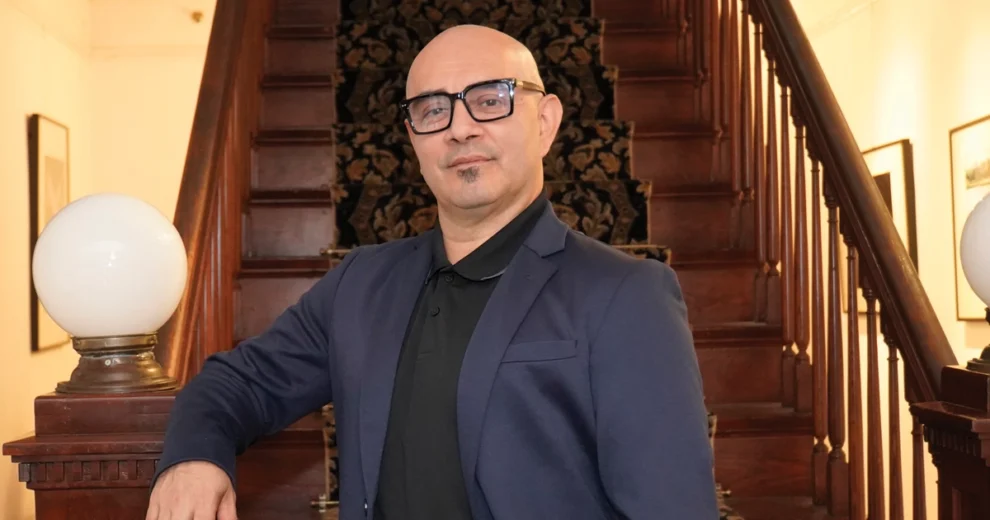 Eriberto Jiménez se encuentra en el interior, con un brazo apoyado en una barandilla de madera pulida frente a una gran escalera. Lleva gafas de montura negra, una chaqueta azul marino y un polo negro.