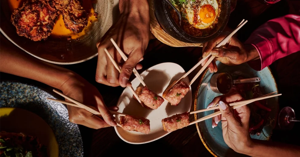 An overhead view at KYU restaurant shows four diners reaching in with chopsticks to share bite-sized appetizers from a central plate.